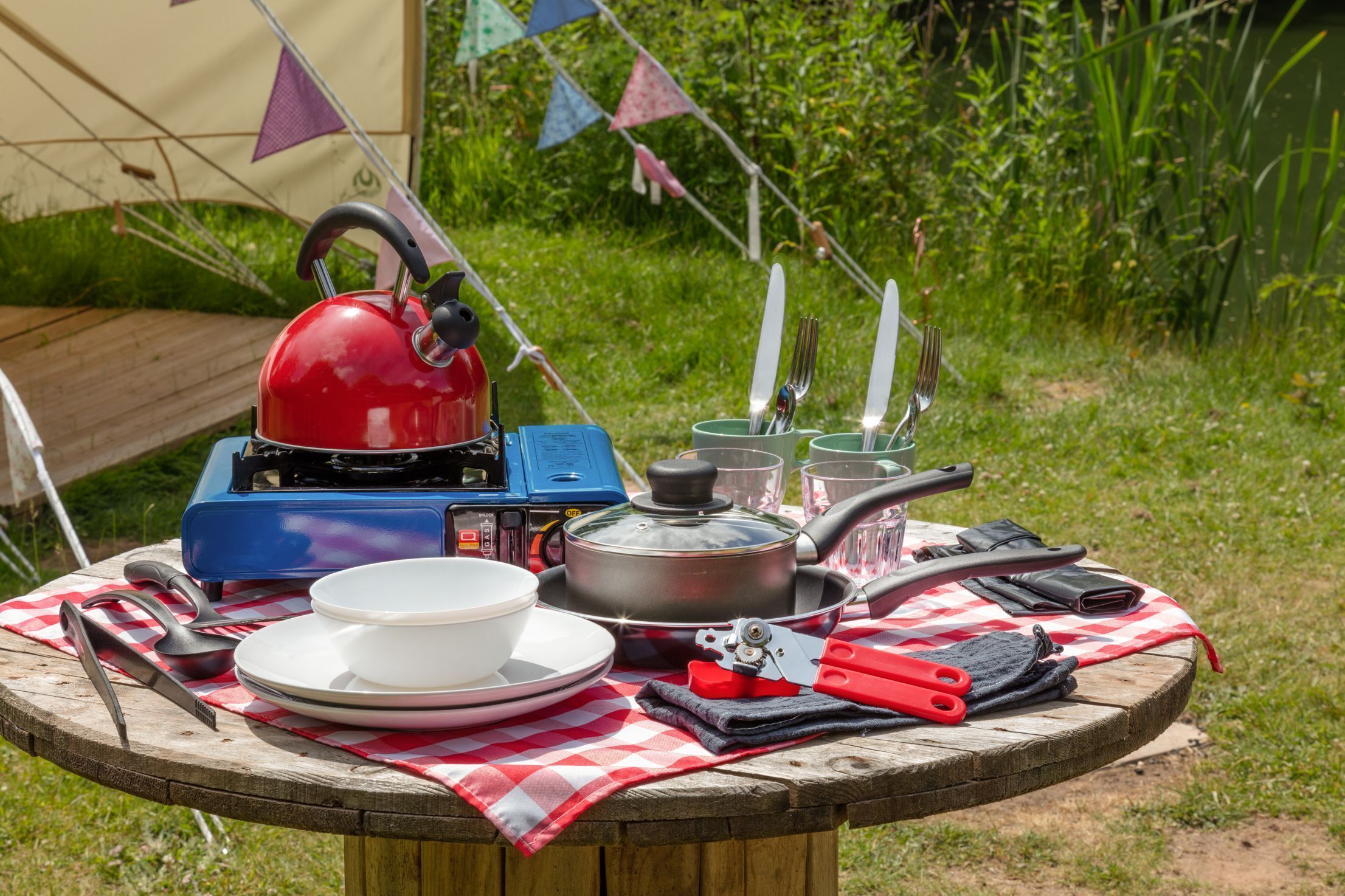 Un coin aménagé pour le camping, avec un réchaud portable, une casserole, des assiettes, des ustensiles de cuisine et une bouilloire rouge posée sur une nappe à carreaux.