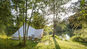 Een tent opgetrokken in een grasrijk gebied, met een tafel en stoelen in de buurt, omgeven door bomen en een waterpartij.