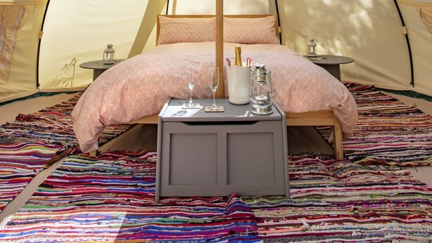 Een tent met een bed, een tafeltje met een fles en glazen, en een kleurrijk vloerkleed.