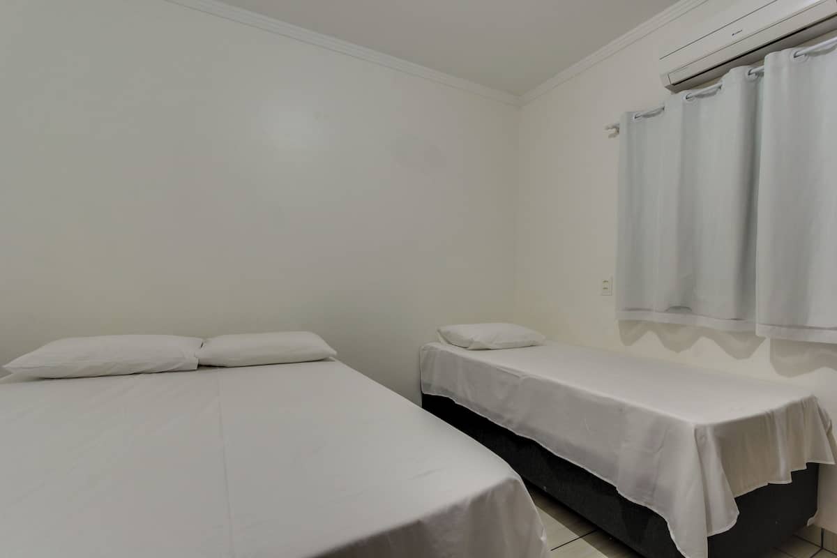 A room with two single beds, white bedding, a window with curtains, and a white air conditioning unit.