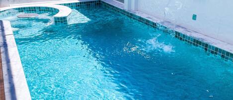 A clear blue swimming pool with a tiled edge, a built-in hot tub, and a white wall with a small window.