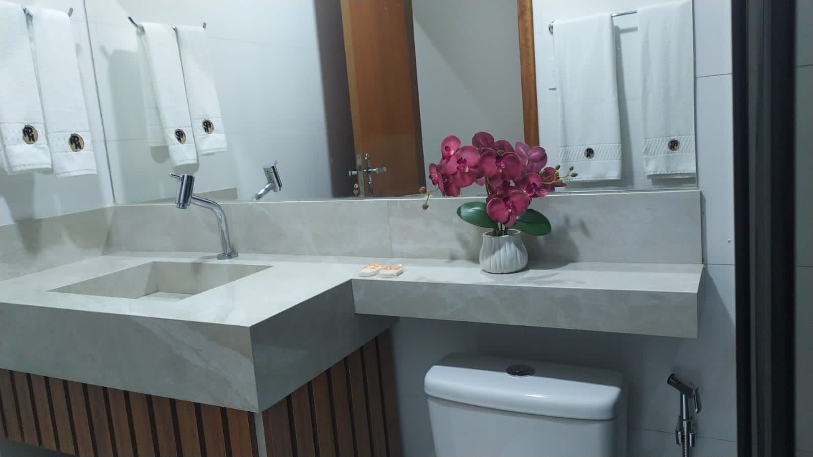 Un baño moderno con un lavamanos grande, un espejo y un inodoro. Hay una florera con flores rosadas sobre la encimera del lavamanos.
