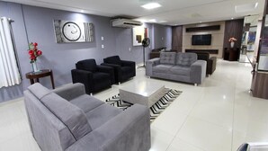 A modern living room with grey sofas, a black armchair, and a grey armchair. There is a flat-screen TV mounted on the wall, a standing fan, and a decorative wall piece. - Augusto Palace Hotel (Maraba)
