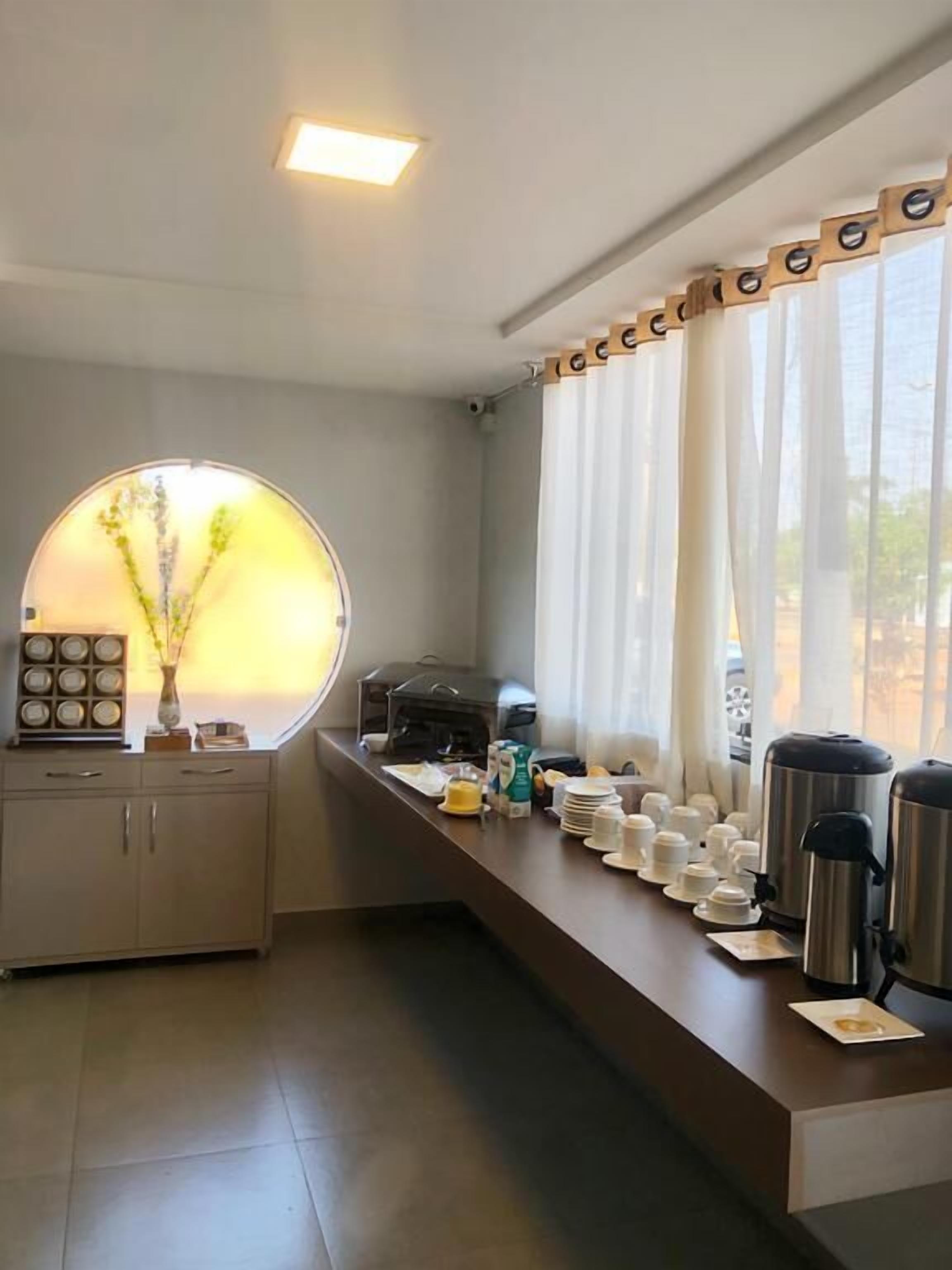 A modern kitchen with a long counter, a coffee machine, and a kettle. There is a window with curtains and a vase with flowers on the counter.