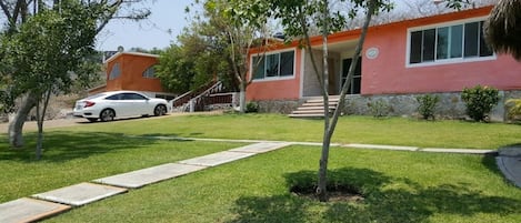 Una propiedad residencial con fachada roja, un coche blanco estacionado en la entrada y un césped cuidado con un sendero que lleva a la puerta.