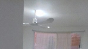 Une petite pièce faiblement éclairée, avec deux lits simples, une fenêtre avec des rideaux et un ventilateur de plafond.