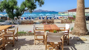 A beachside café with wooden tables and chairs, umbrellas, and a view of the sea and mountains.