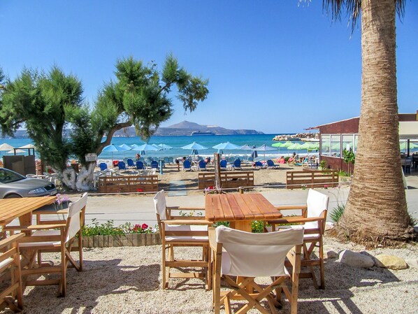 A beachside café with outdoor seating, a palm tree, and a view of the sea and mountains in the background.