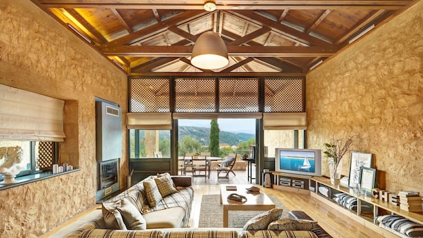 A cozy living room with a sofa, TV, and a fireplace. There are large windows offering a view of the outdoors.