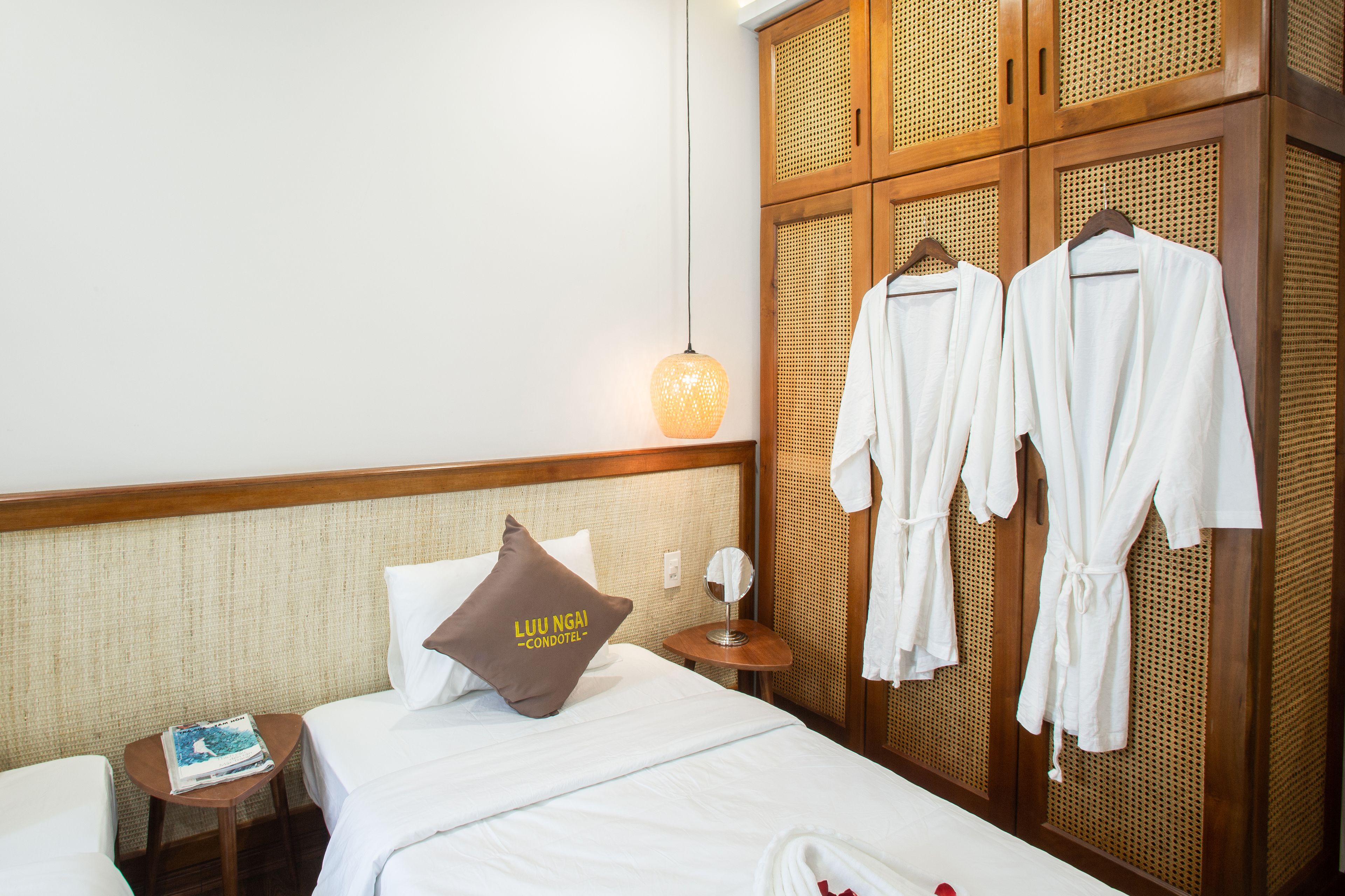 A hotel room with a bed, a bedside table with a book, a lamp, and a wardrobe with robes.