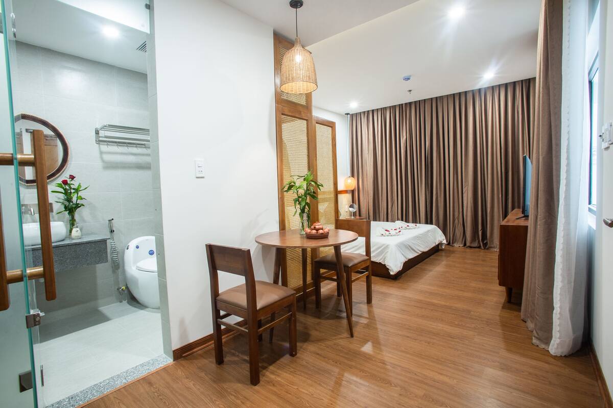 A hotel room with a wooden floor, a round dining table with chairs, a bed with a white bedspread, a bedside table with a lamp, and a large window with curtains.