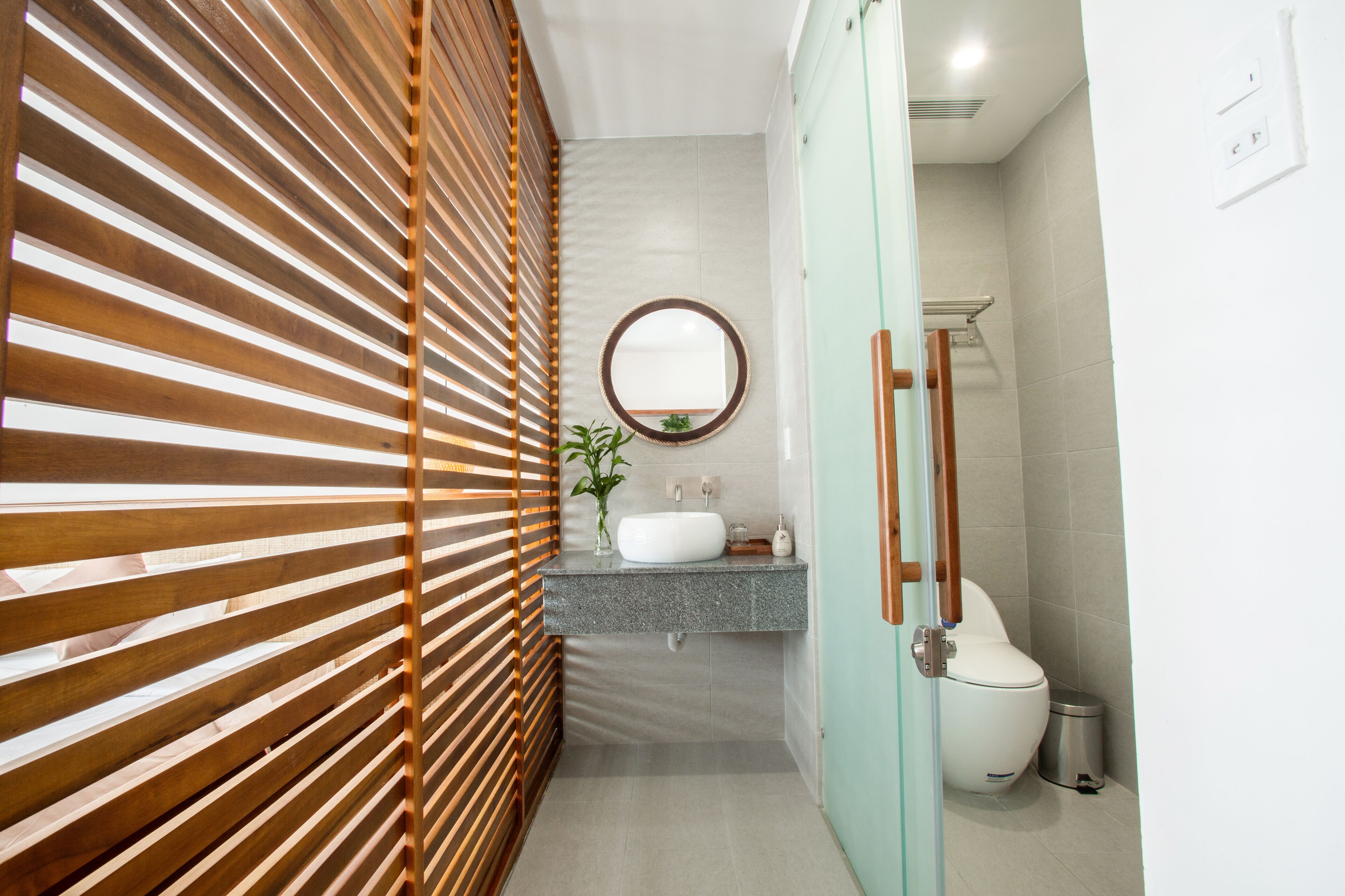 A modern bathroom with a wooden slatted partition, a round mirror, a white sink, and a toilet.