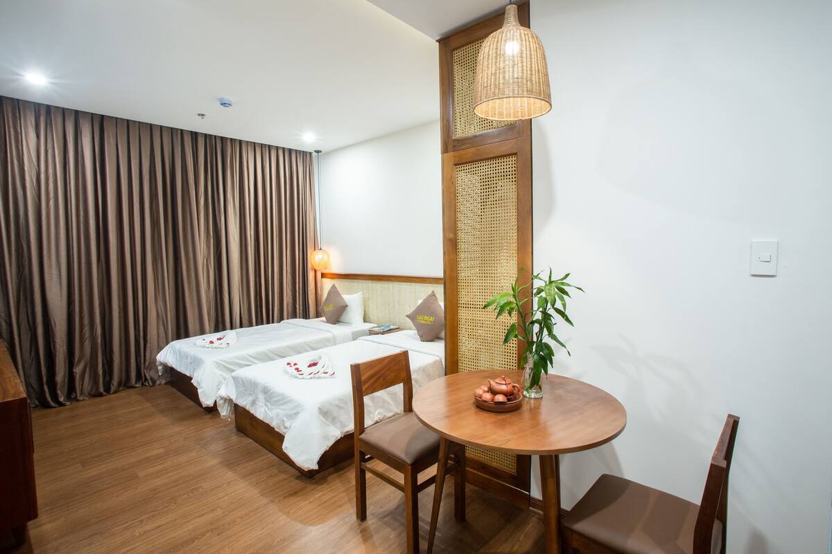 A hotel room with two beds, a dining table with chairs, a hanging light fixture, and a potted plant.