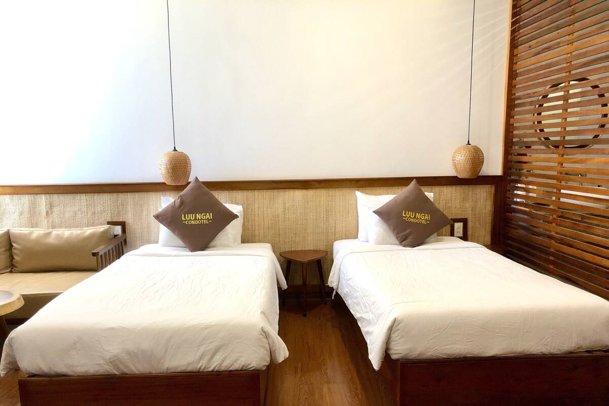 A hotel room with two beds, each with a pillow labeled "LUU NGAI CONDOTEL". There is a small round table between the beds, and the room has wooden flooring and a wooden headboard with slats.