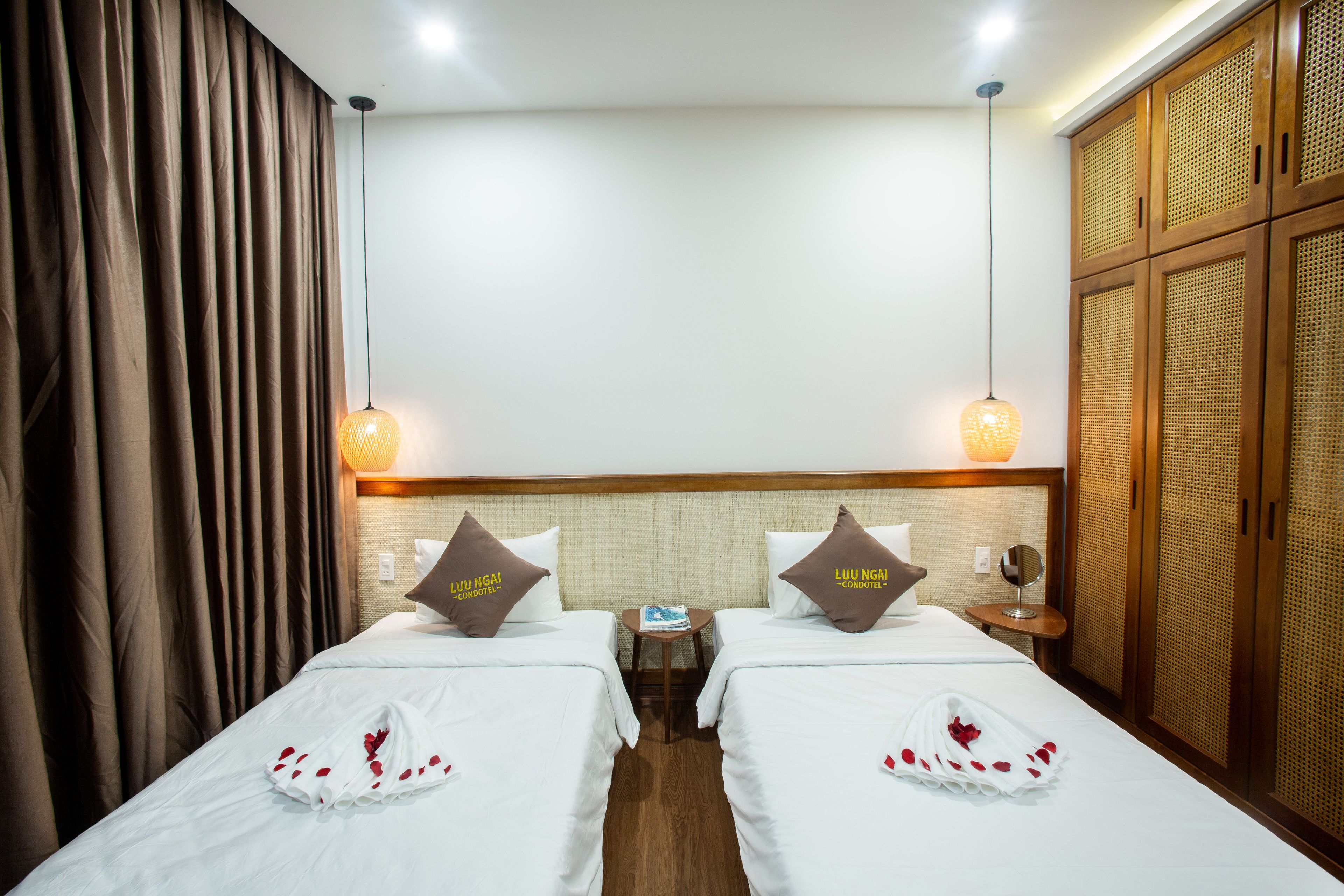 A hotel room with two beds, each with a pillow labeled "Luu Ngai Condotel". There is a small table between the beds, a mirror on the wall, and a wardrobe with a woven design.
