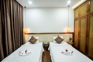 A hotel room with two beds, each with a pillow labeled "Luu Ngai Condotel". There is a small table between the beds, a mirror on the wall, and a wardrobe with a woven design.