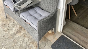 Un canapé en osier, avec un ordinateur portable, des coussins gris et une porte vitrée donnant sur la cuisine.