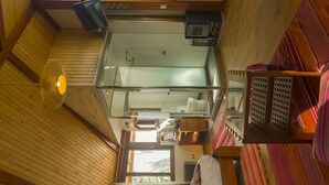Una habitación con techos altos, paredes de madera, un balcón con cerramiento de vidrio, una pequeña cocina y un ventanal que proporciona luz natural.
