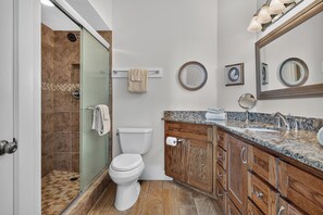 Ein Badezimmer mit Glasdusche, Toilette, Holzschränken, einer Granitarbeitsplatte und einem großen Spiegel.