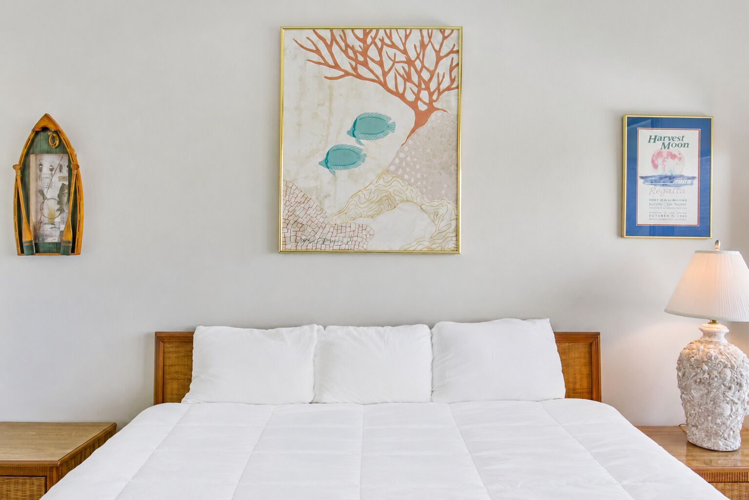 A neatly made bed with white linens, a wall-mounted painting of a tree with fish, a framed certificate, and a lamp on a nightstand.