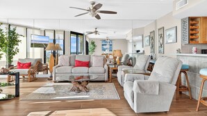 Un salón amplio con un gran ventanal, ventilador de techo y una zona de cocina con armarios de madera.