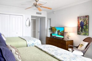 A hotel room with two beds, a television, a ceiling fan, a wooden dresser, and a wall painting. - Sandpiper Condominiums 605, Panoramic Gulf Views w/ direct access to Mustang Island/Port A beaches. (Port Aransas)