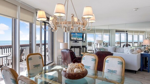A spacious living room with a glass dining table, comfortable seating, a large chandelier, and a view of the ocean.