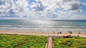A beach with a wooden boardwalk leading to the shore, a sandy beach, and a clear blue sky with clouds.