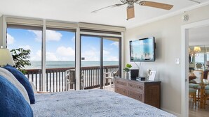 A bedroom with a large bed, a ceiling fan, a TV, and a view of the ocean through large sliding doors.