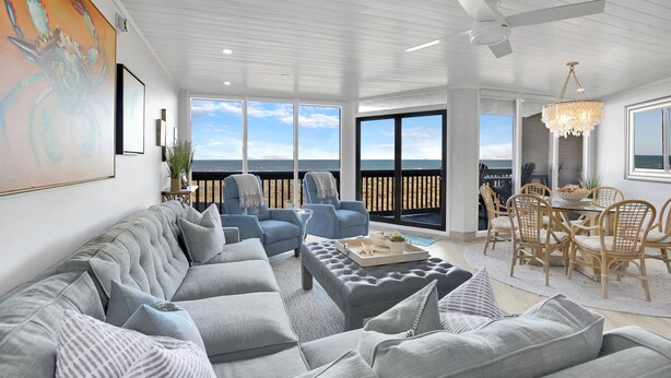 A modern living room with a large gray sectional sofa, a dining area with wooden chairs, and a view of the ocean through large windows.