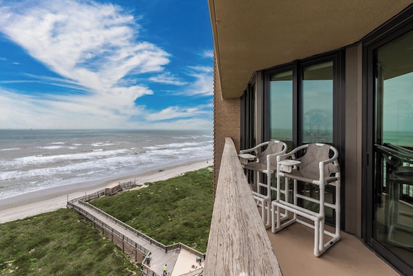 A balcony with a view of the beach and ocean, a white chair, and a glass door.