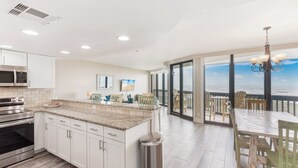 Una cocina moderna con armarios blancos, encimeras de granito y electrodomésticos de acero inoxidable. Cuenta con una isla grande con un lavamanos, un comedor con sillas de madera y vistas al océano a través de amplias ventanas.