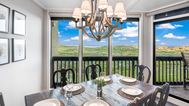A dining area with a table set for six, a chandelier, and large windows offering a view of a hilly landscape.