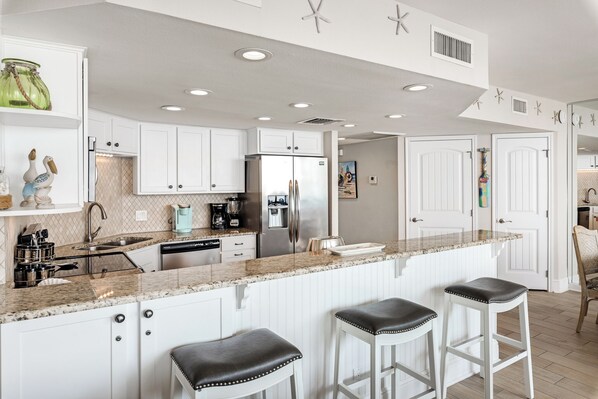 Une cuisine moderne avec des meubles blancs, un grand îlot central doté de tabourets, des appareils en acier inoxydable et des éléments décoratifs sur les murs.