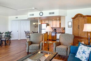 A spacious living room with two patterned armchairs, a coffee table, and a blue sofa with star pillows. A dining area with a table and chairs is visible in the background. - Sandpiper Condominiums 1007L, Panoramic Gulf View w/ direct access to Mustang Island/Port A beaches. (Port Aransas)