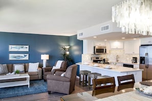 Ein modernes Wohnzimmer mit einer Couch, Sesseln und einem Couchtisch. Im Hintergrund ist eine Küche mit Mikrowelle, Herd und Kühlschrank zu sehen.