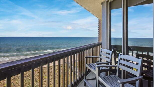 Un balcon avec deux chaises en bois donnant sur une plage et l’océan.