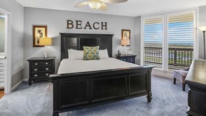 Une chambre avec un grand lit, un ventilateur de plafond et une vue sur la plage par la fenêtre.