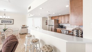 Une cuisine moderne avec des comptoirs blancs, des appareils en acier inoxydable et des armoires en bois. On y trouve trois tabourets de bar blancs avec des coussins à motifs.