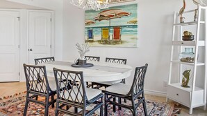 Un comedor con una mesa redonda, sillas negras, una araña y un cuadro con motivos playeros en la pared.