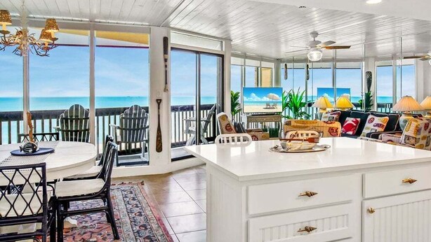 Amplia habitación con un gran ventanal con vistas al océano, una encimera blanca tipo isla y un comedor con sillas y mesa.