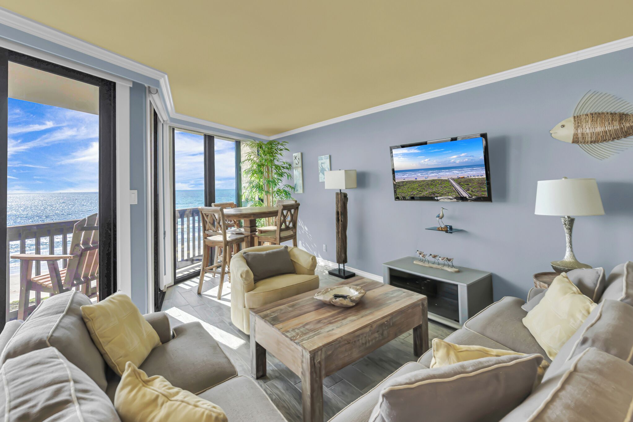 Ein Wohnzimmer mit großem Fenster zum Meer, einem an der Wand montierten Fernseher und einem Holztisch.