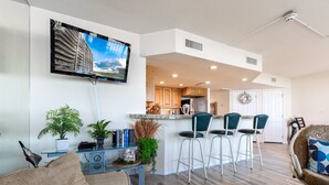 A modern living room with a flat-screen TV, a kitchen with bar stools, and a dining area. - Sandpiper Condominiums 303, Panoramic Gulf Views w/ direct access to Mustang Island/Port A beaches. (Port Aransas)