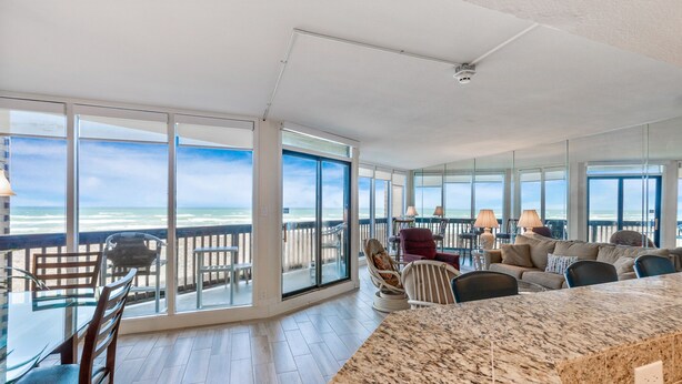 A spacious living area with a large glass wall offering a view of the ocean. There is a dining table with chairs, a sofa, and a coffee table. - Sandpiper Condominiums 303, Panoramic Gulf Views w/ direct access to Mustang Island/Port A beaches. (Port Aransas)