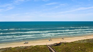 Une plage avec une promenade en bois menant à une étendue de sable, quelques personnes et un véhicule garé près de l’eau.