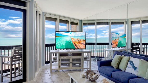 Een balkon met uitzicht op zee, een televisie op een standaard, een blauwe bank en een houten salontafel.