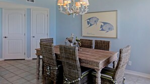 Een eetkamer met een houten tafel en rieten stoelen, een kroonluchter en een schilderij van een vis aan de muur.