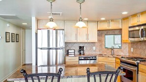 A modern kitchen with stainless steel appliances, wooden cabinets, granite countertops, and two bar stools. - Sandpiper  #1007: Beachfront 2 Bedroom 2 Bathroom With Spectacular Views and 24 Hour Management (Port Aransas)