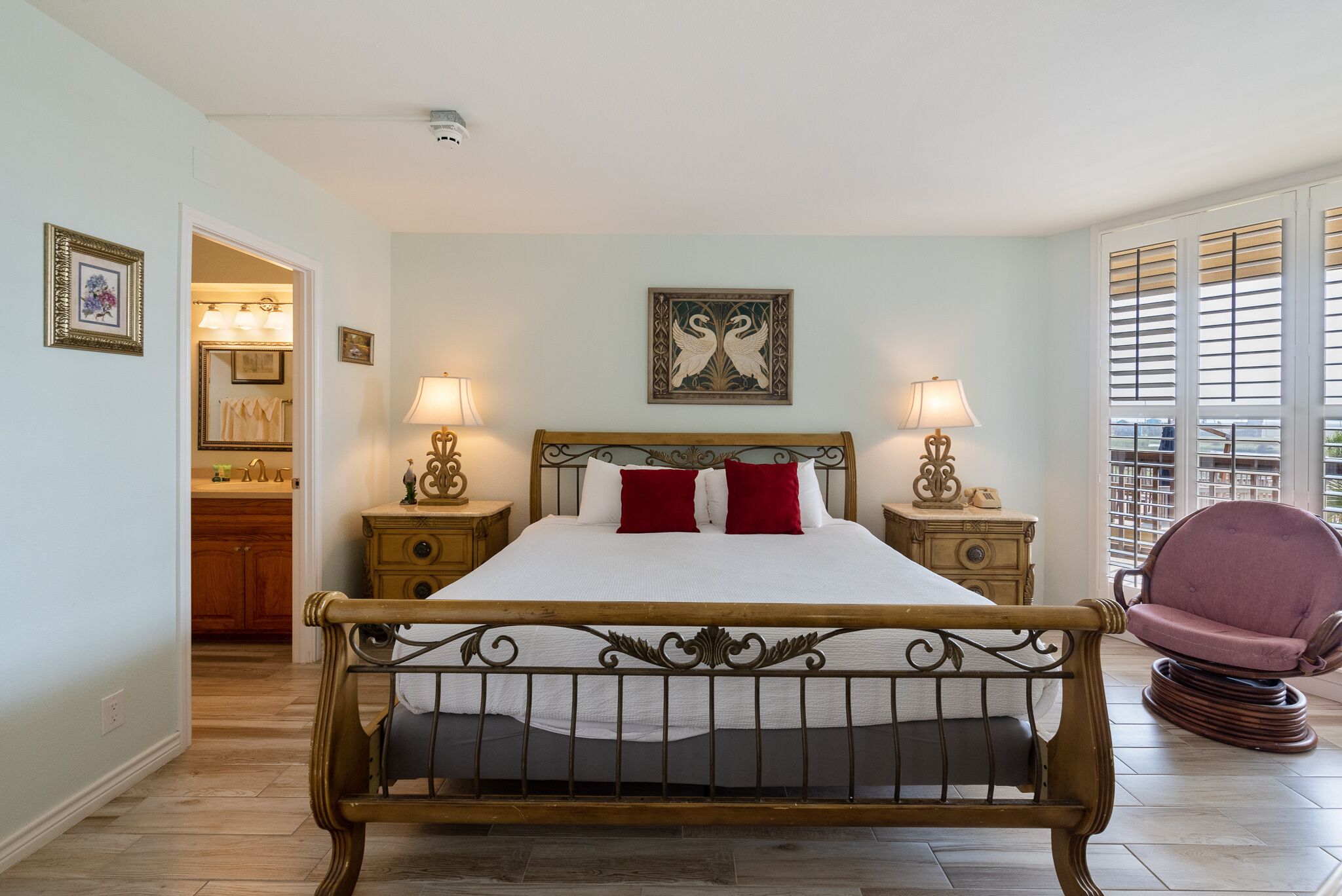 A bedroom with a wooden bed, two bedside tables with lamps, a chair, and a view of the outside through large windows.