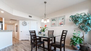 A dining area with a glass table, wooden chairs, a chandelier, and potted plants. - Sandpiper Condominiums 303L, Panoramic Gulf Views w/ direct access to Mustang Island/Port A beaches. (Port Aransas)
